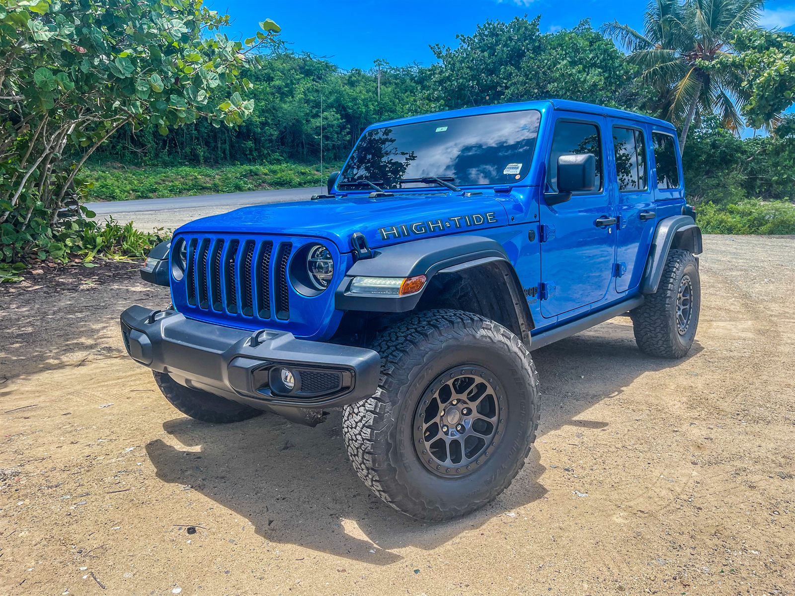 Blue Jeep Wrangler High Tide parked on a sandy track with palms and sea grape behind