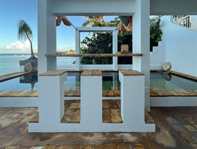 Shaded outdoor bar with a timber counter and stools, looking across the cocktail pool to the sea