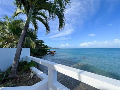 White balcony railing framed by a palm, looking along the coastline over clear shallow water