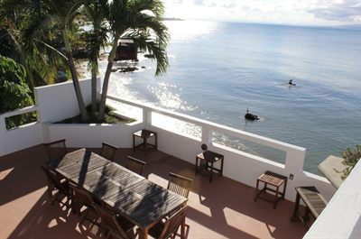 Terrace with a long teak table above the water, palms to one side and the sea catching early light
