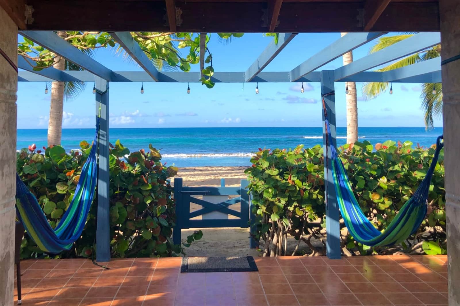 Covered patio with two hammocks under a blue pergola, looking through a garden gate to the turquoise Caribbean